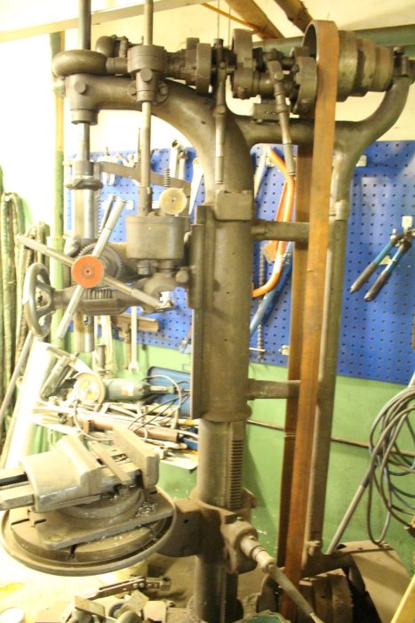 En gammal pelarborrmaskin i ett verktygsfyllt garage med synlig drivrem och borrchuck.