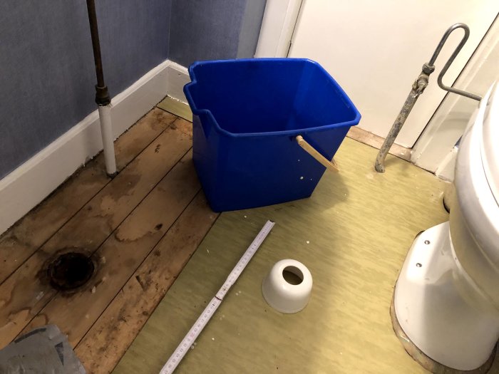 Läckage i ett badrum med en synlig vattenskada på golvet vid gammalt rör, toalett och en hink.