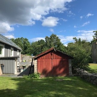 Röd träbod i en trädgård med grönt gräs, stenmur och del av hus i bakgrunden under blå himmel.