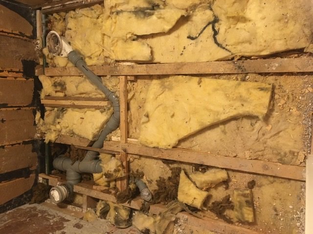 Delvis isolerad vägg med exponerad rördragning och sliten gul isolering mellan träreglar.