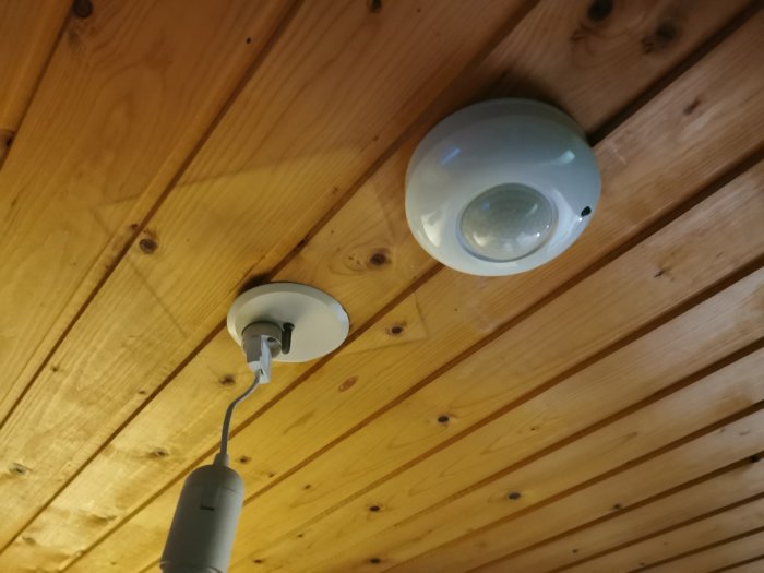 Rörelsesensor monterad i taket nära en lampa på träpanelbakgrund.