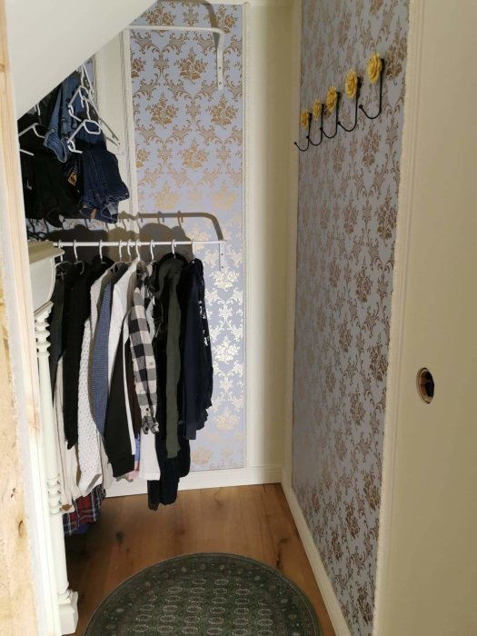 Garderob under trappa med hängande kläder och väggkrokar mot tapetserad vägg.