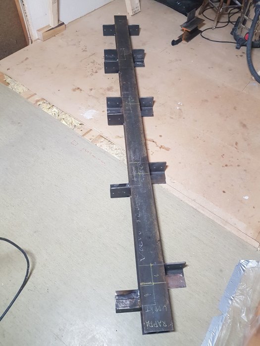 Steel beam with welded brackets on a wooden floor, featuring chalk markings and nearby construction tools.