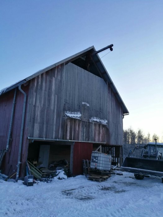 Vintervy av en stor röd lada med en höhiss och diverse byggmaterial framför, mot en klar himmel.