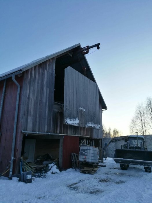 Gammal höhiss i en röd ladugård som lyfts ned med hjälp av en grävmaskin, omgiven av snö och byggbråte.