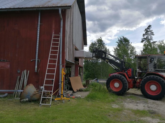 En traktor med lyftarm vid en röd lada, med byggmaterial och en stege på marken, symboliserar landsbygdsrenovering.