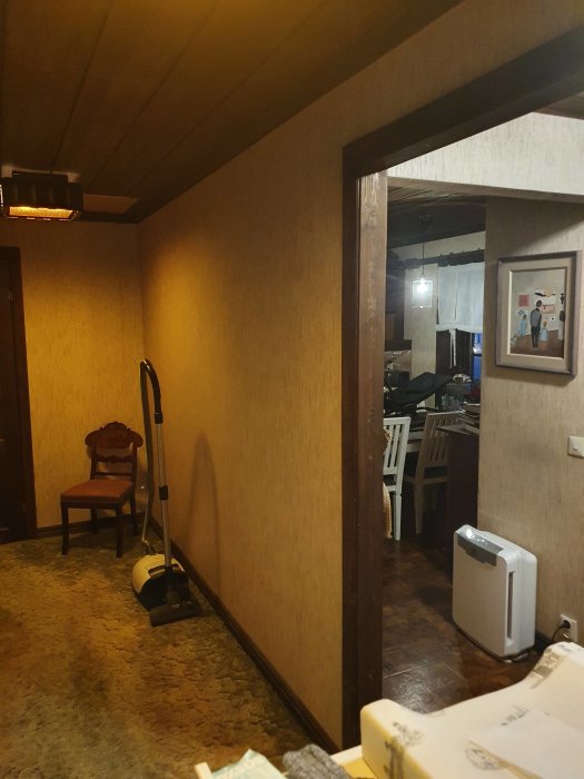 Talon käytävä, jossa villamatto ja puupaneelit, näkyy avoin oviaukko keittiöön. Oikealla nojatuoli ja pölynimuri.