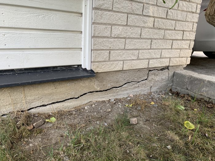 Horisontell spricka i bottenplatta nära hörnet på ett hus, med synlig fukt och skadad mark omkring.