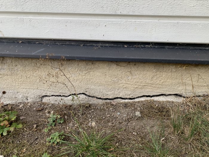 Horisontell spricka längs bottenplattan vid ett hörn av ett hus, omgivet av gräs och jord.