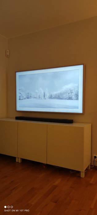 Installerad tv på väggen ovanför en vit byrå med en ansluten soundbar och en vinterbild på skärmen.