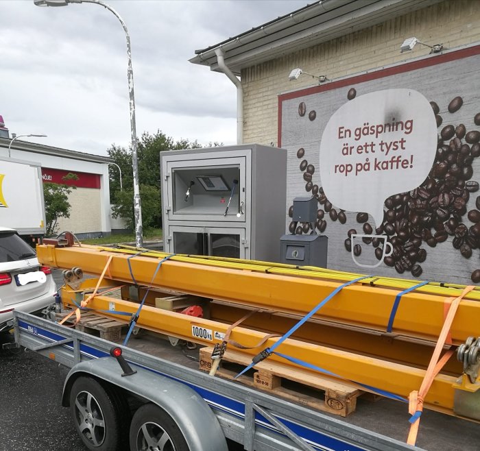 Gul traversbalk lastad på släpvagn framför reklamskylt med texten "En gäspning är ett tyst rop på kaffe".