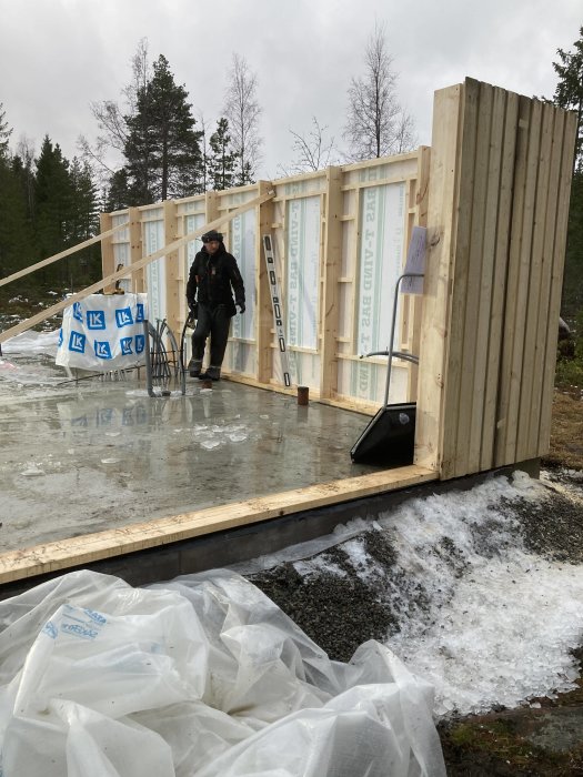 Byggplats med uppresade träväggar och bärlinor på en betongplatta, person i byggkläder och smältande snö runtomkring.