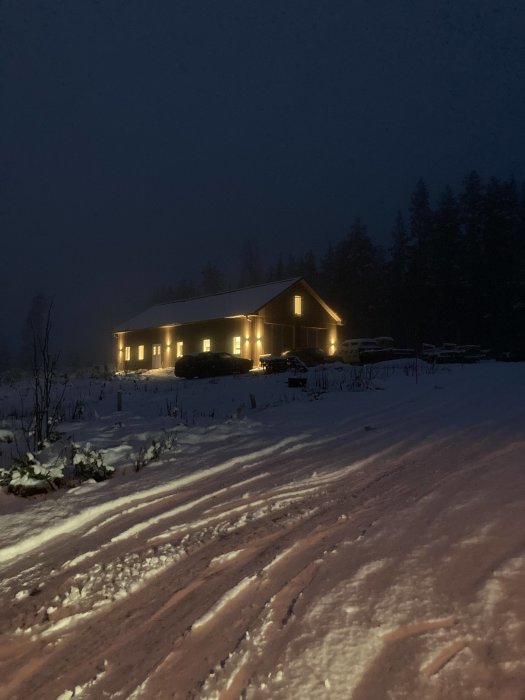 Kvällsbild av ett hus med tända lampor och nysatt fönster på gaveln, omgivet av snötäckt mark.