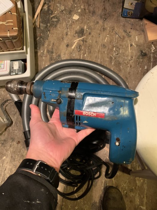 Hand håller en gammal sliten blå Bosch borr med träspån i bakgrunden, synbar slitage och fokuserad på växelknappen nära chucken.