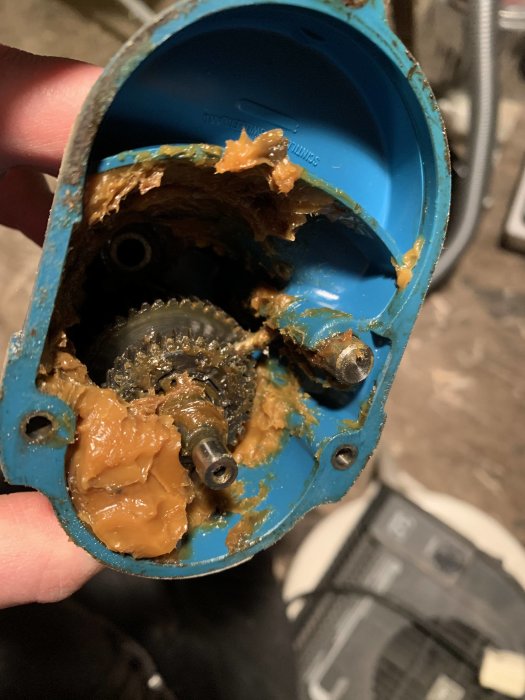 Interiör av en äldre maskin med blå kåpa och brunsmetigt kugghjul, täckt med fett, under reparation.