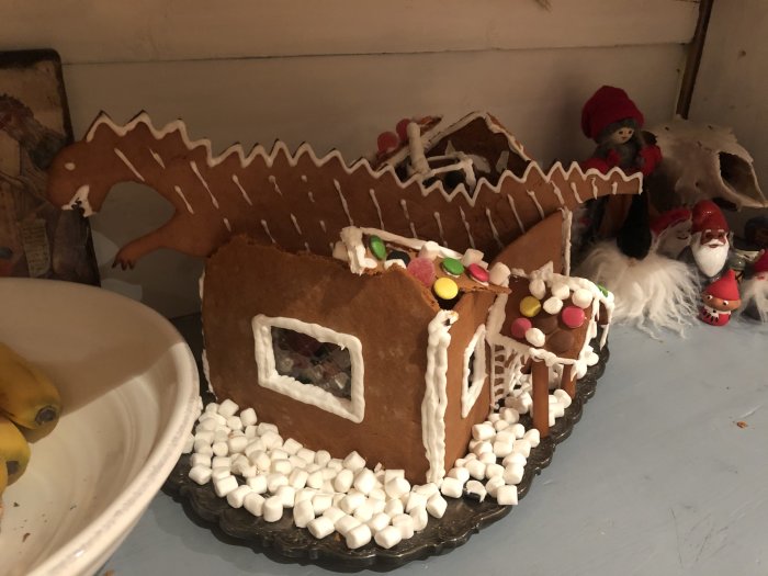 Pepparkakshus med kollapsat tak och en pepparkaksdinosaurie i bakgrunden, omgivet av juldekorationer.