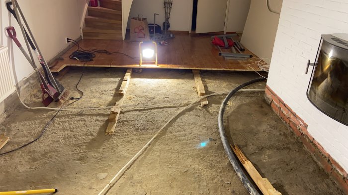 Uppbrutet golv i gillestuga med synlig elslang och reglar, bredvid en kamin och arbetslampa.