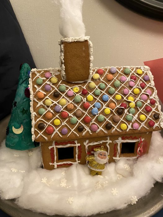 Handgjord pepparkakshus dekorerad med färgglatt godis och glasyr, en liten gran och en julfigur bredvid på ett vitt bomullstäcke.