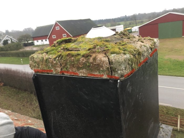 Mossbeklädd skorstenskrag med markerade sprickor, bakgrund med lantlig miljö och byggnader.