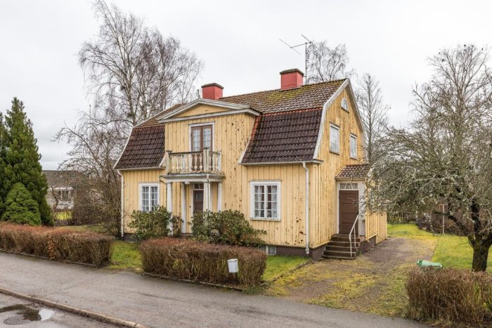 Ett gulmålat trähus med tegeltak som behöver renoveras, omgivet av en trädgård och kala träd.