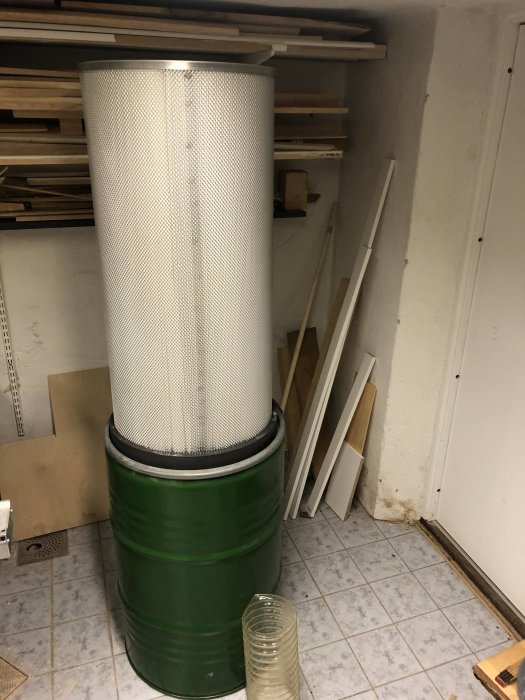 Grön tunna med stort cylindriskt filter ovanpå i ett förrådsutrymme med trämaterial i bakgrunden.