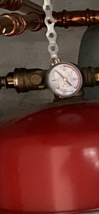 Tryckmätare på kopparledning ovanför röd expansionskärl med bakgrund av mörkgrå vägg.