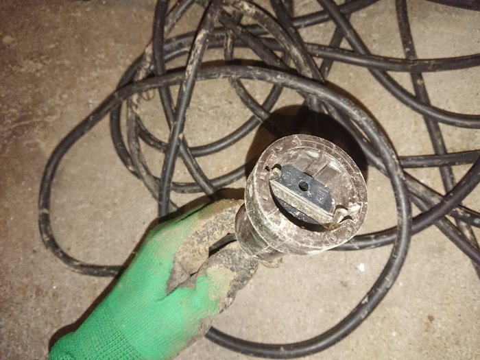 Ett lerigt och slitet jordat eluttag i änden av en förlängningssladd hålls upp framför en hög av sladdar.