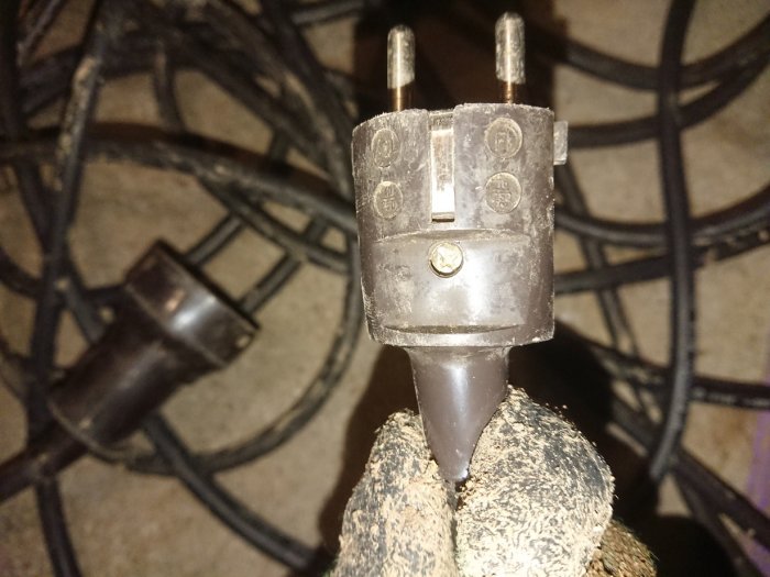 Jordad skarvsladd i närbild hållen av hand med skyddshandske, bakgrunden visar en röra av sladdar.