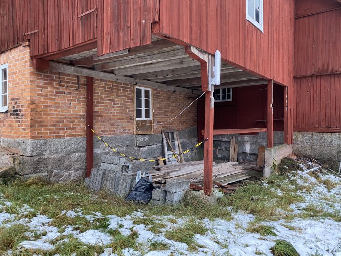 Outnyttjat hörn under rödmålad byggnad med tegel och trädetaljer, betongfundament och lite snö på marken.