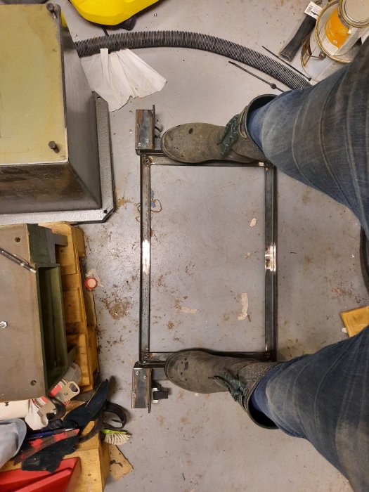 Person står ovanför en svetsad metallram på ett verkstadsgolv med verktyg omkring.