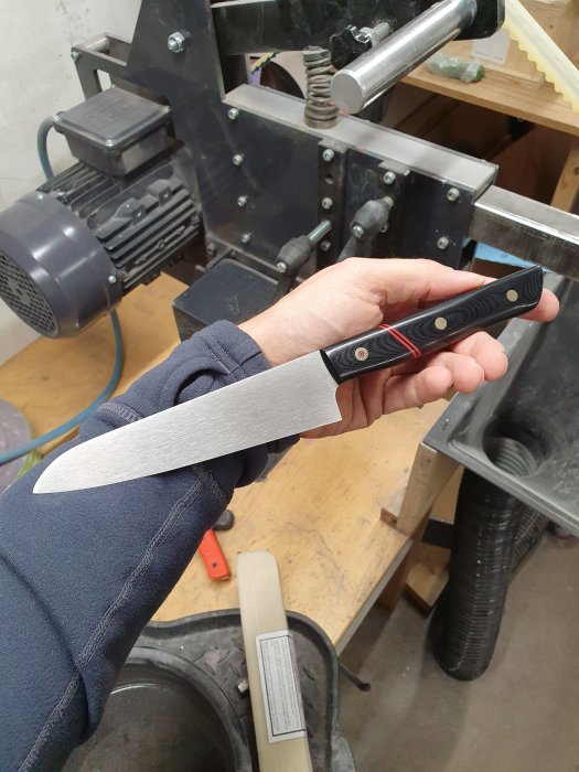 Hand håller en nyslipad kniv med svart handtag i en verkstad.