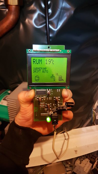 En hand håller ett elektroniskt kretskort med en skärm som visar temperatur och värmepumpstatus.