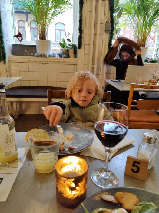 Barn som äter lunch vid ett restaurangbord med glas vin och levande ljus i förgrunden.