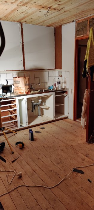 Kök under renovering med borttagna skåp, synliga vattenrör och avlopp, och arbetsmaterial på ett trägolv.