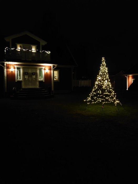 Ljusgran med vita lampor lyser upp natt i trädgård framför hus med julbelysning på balkongen.