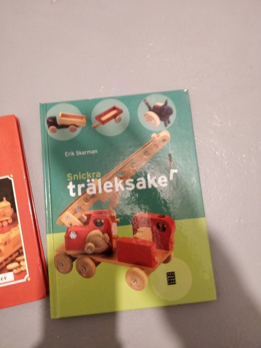 Bokomslag 'Snickra träleksaker' med bilder på handgjorda träleksaker, placerad bredvid en röd bok.