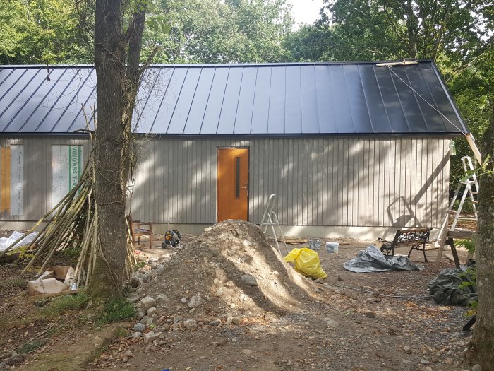 Hus under renovering med ojämnt grus och byggmaterial framför fasaden i höstsol.