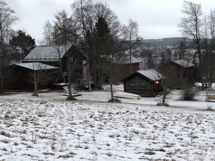 Vinterlandskap med byggnader; till vänster en utbyggnad och garage, vedbod och loftbod till höger.