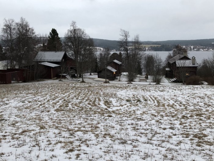 Utsikt över ett snötäckt landskap med flera byggnader inklusive ett hus, garage, vedbod och lofthus, vid en sjö.