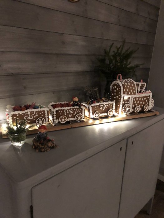 Ett pepparkakståg dekorerat med glasyr och godis står på en byrå med tända ljus och juldekorationer i bakgrunden.