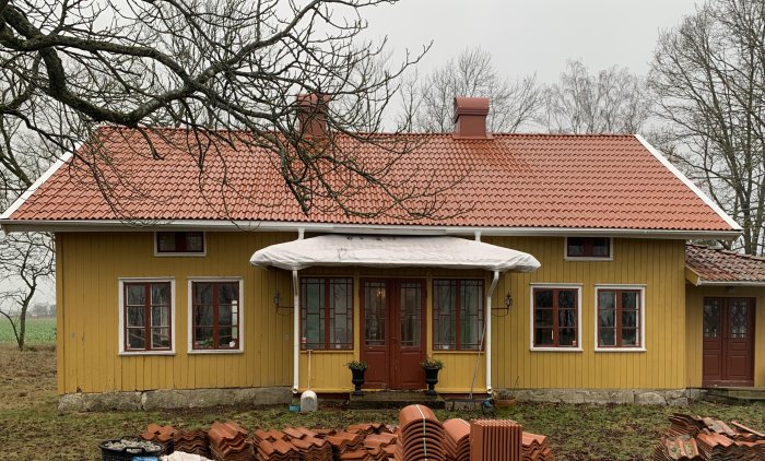 Gult hus med nytt rött tegeltak, vita fönsterknutar och tillhörande gamla taktegel samlade på marken.