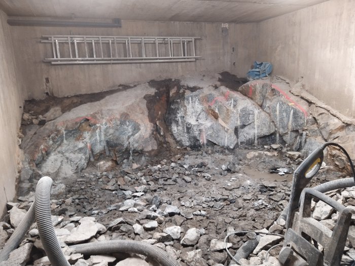 Delvis sprängd bergdel med stora stenblock och sprickor, märkta med rödfärg, i en byggplats med betongväggar och grus.