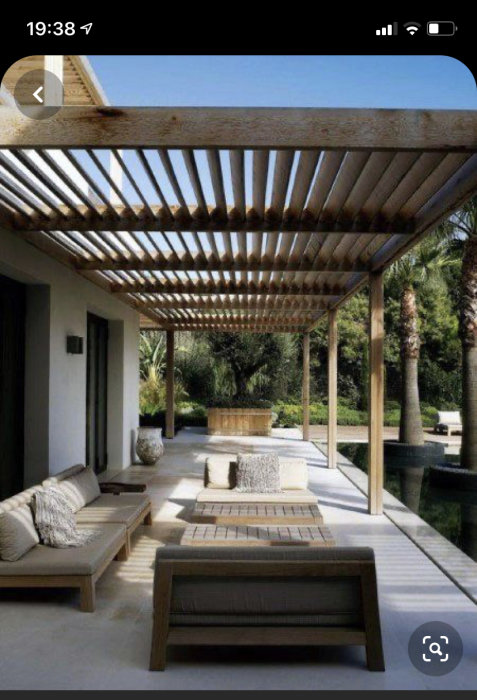 Träpergola med parallella takbjälkar över betongaltan, omgiven av trädgård och möbler.