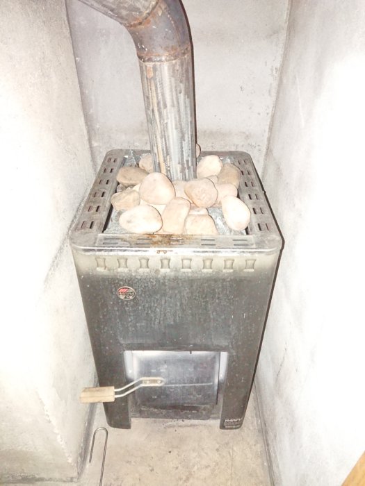 En bastuugn belagd med stenar i ett hörn, med synlig skorsten och överbelyst av kamerans blixt.