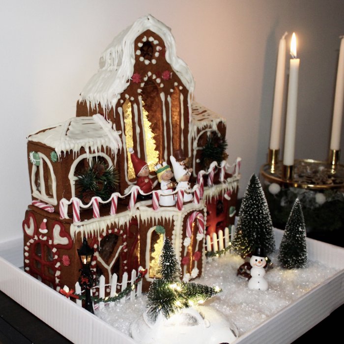 Komplex pepparkakskreation av ett hus dekorerat med godis, glasyr och figurer av tomtenissar omringat av små granar.