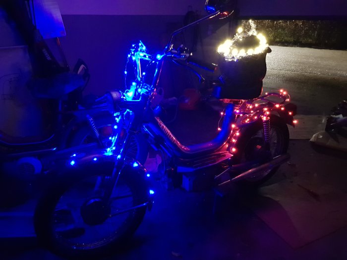 En moped dekorerad med blå och röda ljusslingor i ett mörkt garage, redo för luciatåg.