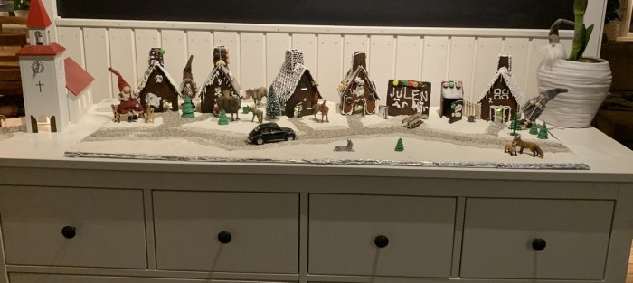 Familjebyggda pepparkakshus står på en byrå med mjöl som snö, dekorerad med små figurer och en kyrka, i en julig miniatyrby.