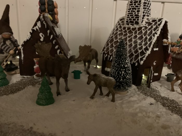 Familjegjorda pepparkakshus med glasyrvindlingar, omgivna av renfigurer och julgranar i ett vinterlandskap av mjöl.