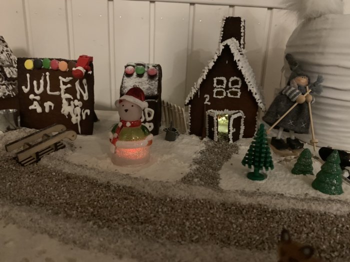 Fem hemgjorda pepparkakshus av olika storlekar med juldekorationer och en snögubbe omgiven av mjölsnö och miniatyrplastgranar.