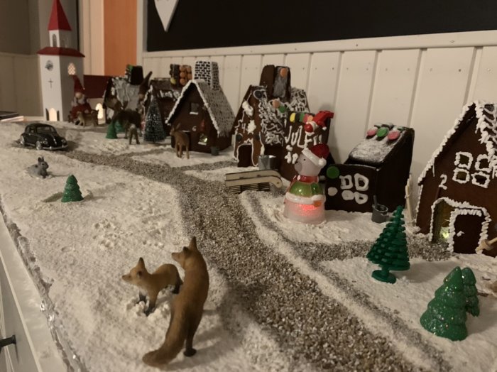 Ett arrangemang av handgjorda pepparkakshus med ett vinterlandskap av mjöl, små djurfigurer och ett juldekorerat bilmodell.